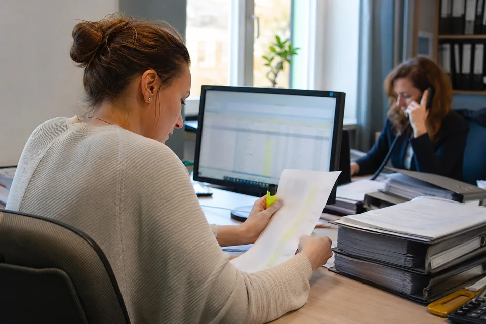 Księgowa porównuje wydruk długiej listy kontrahentów z arkuszem na monitorze